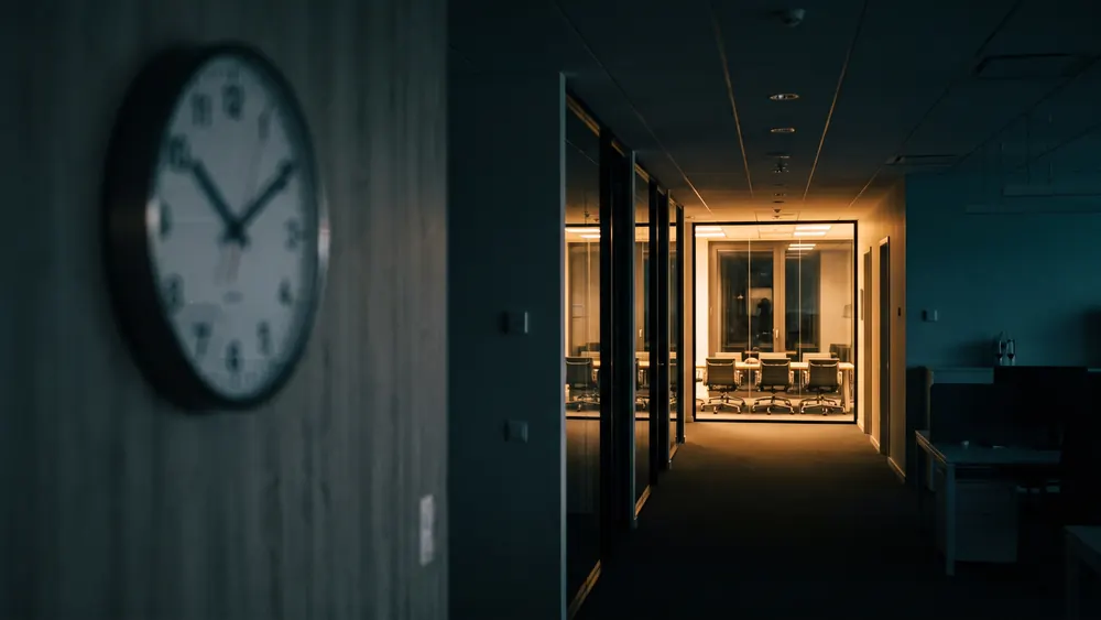 Bürokorridor am späten Abend, ein Glasraum noch beleuchtet, der Rest liegt dunkel, im Vordergrund unscharf eine Wanduhr