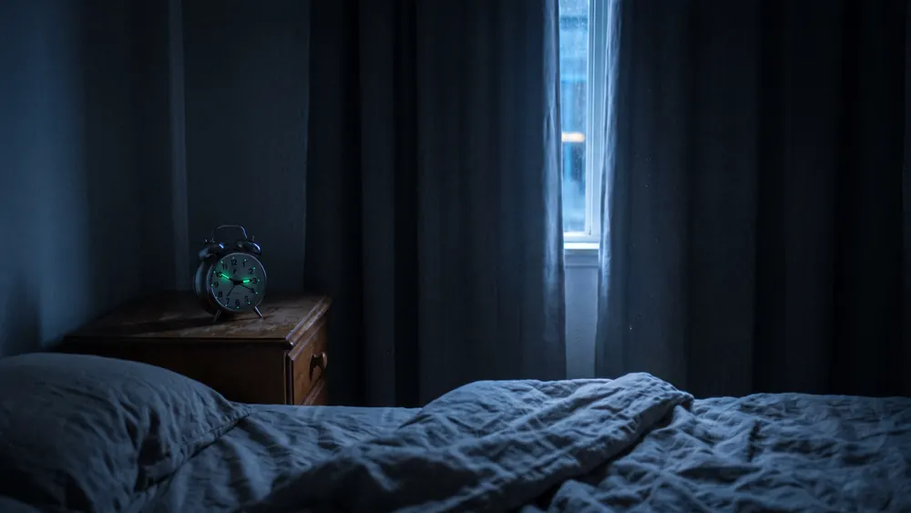 Dunkles Schlafzimmer mitten in der Nacht, ein analoger Wecker auf dem Nachttisch zeigt kurz nach drei, schwaches Straßenlicht am Vorhangspalt